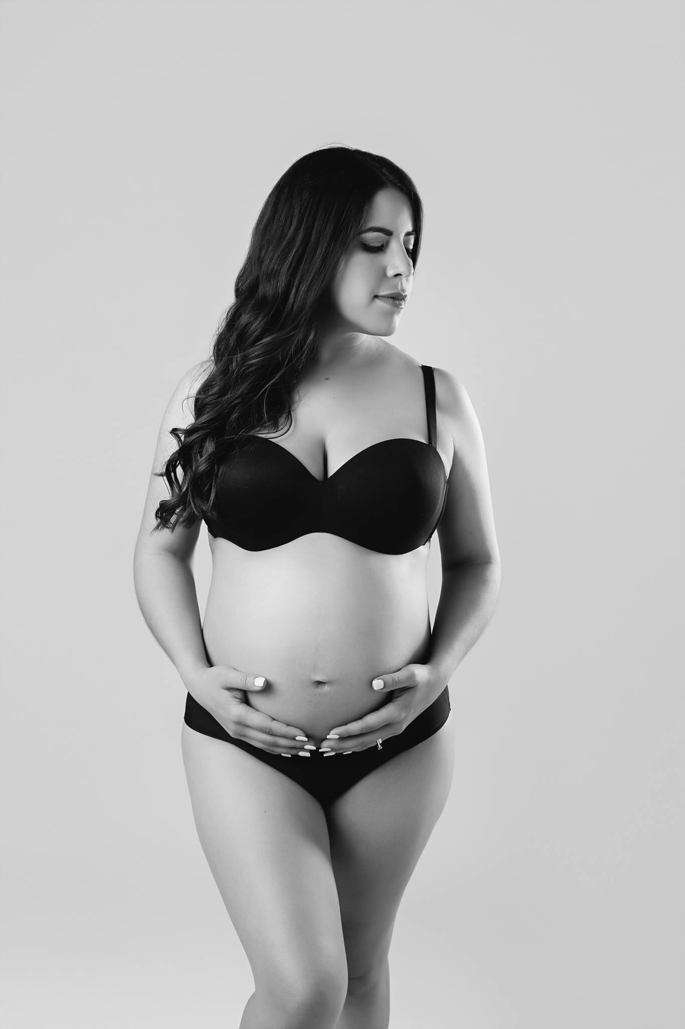 A pregnant woman in black underwear stands in a studio holding her bump and looking down her shoulder