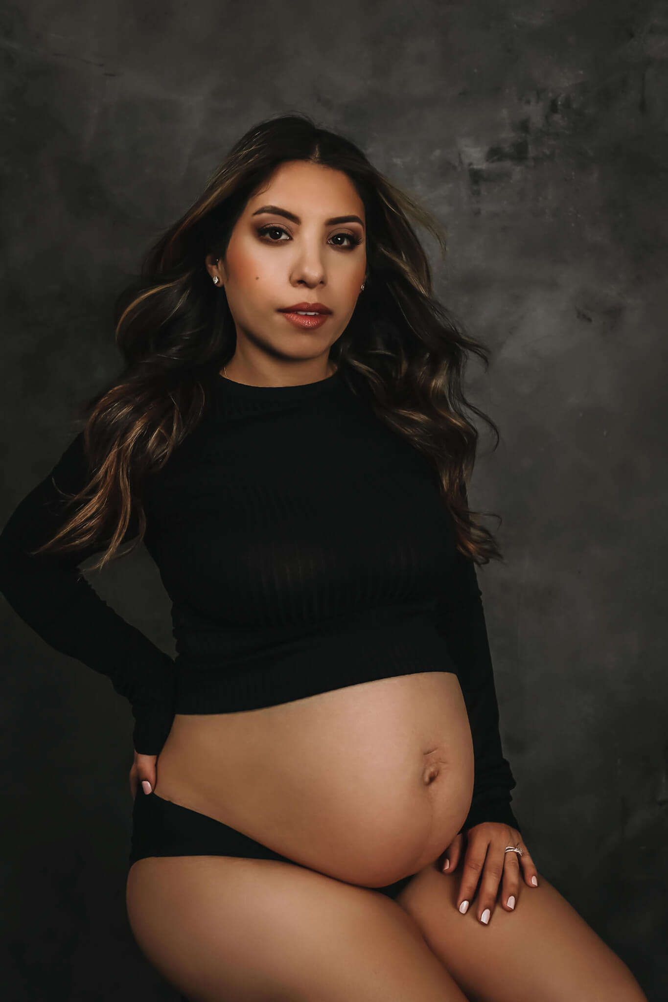 A woman in black underwear and top sits on a stool in a studio with bump exposed after meeting austin midwives