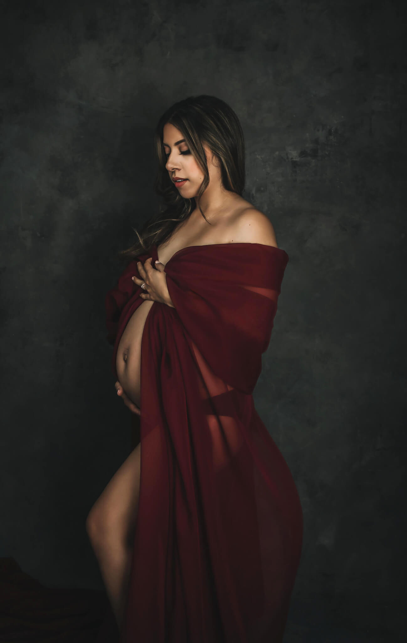 A mother to be stands in a studio covering with a long red fabric and holding her exposed bump after meeting austin midwives