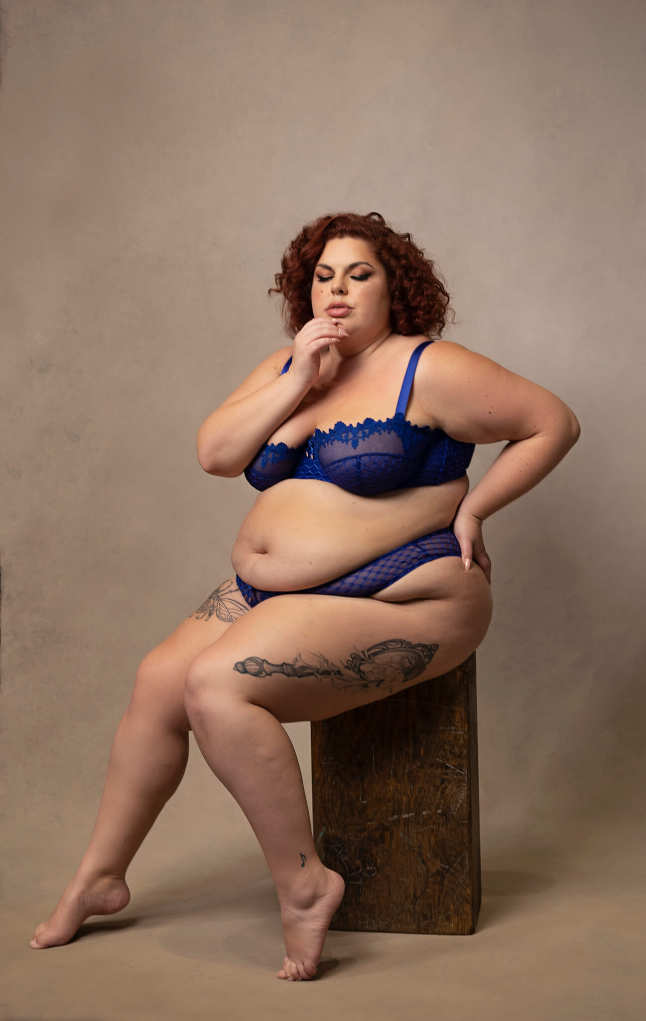 A woman in bright blue lingerie sits on a wooden block in a studio with a hand on her chin after using austin nail salons
