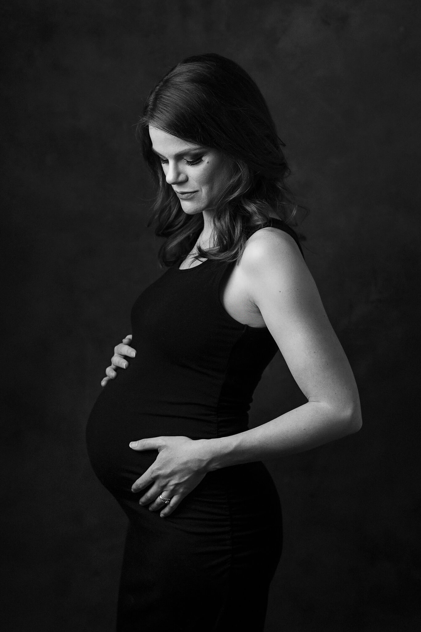 A mother to be stands in a studio smiling down at her bump in her hands in a black dress after a 3d ultrasound austin