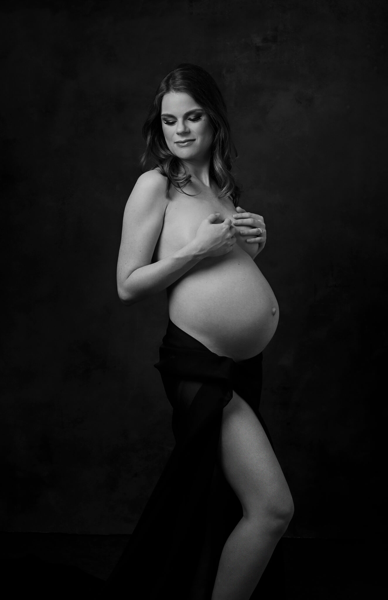 A B&W photo of a pregnant woman standing in a studio with no shirt and smiling down her shoulder after a 3d ultrasound austin