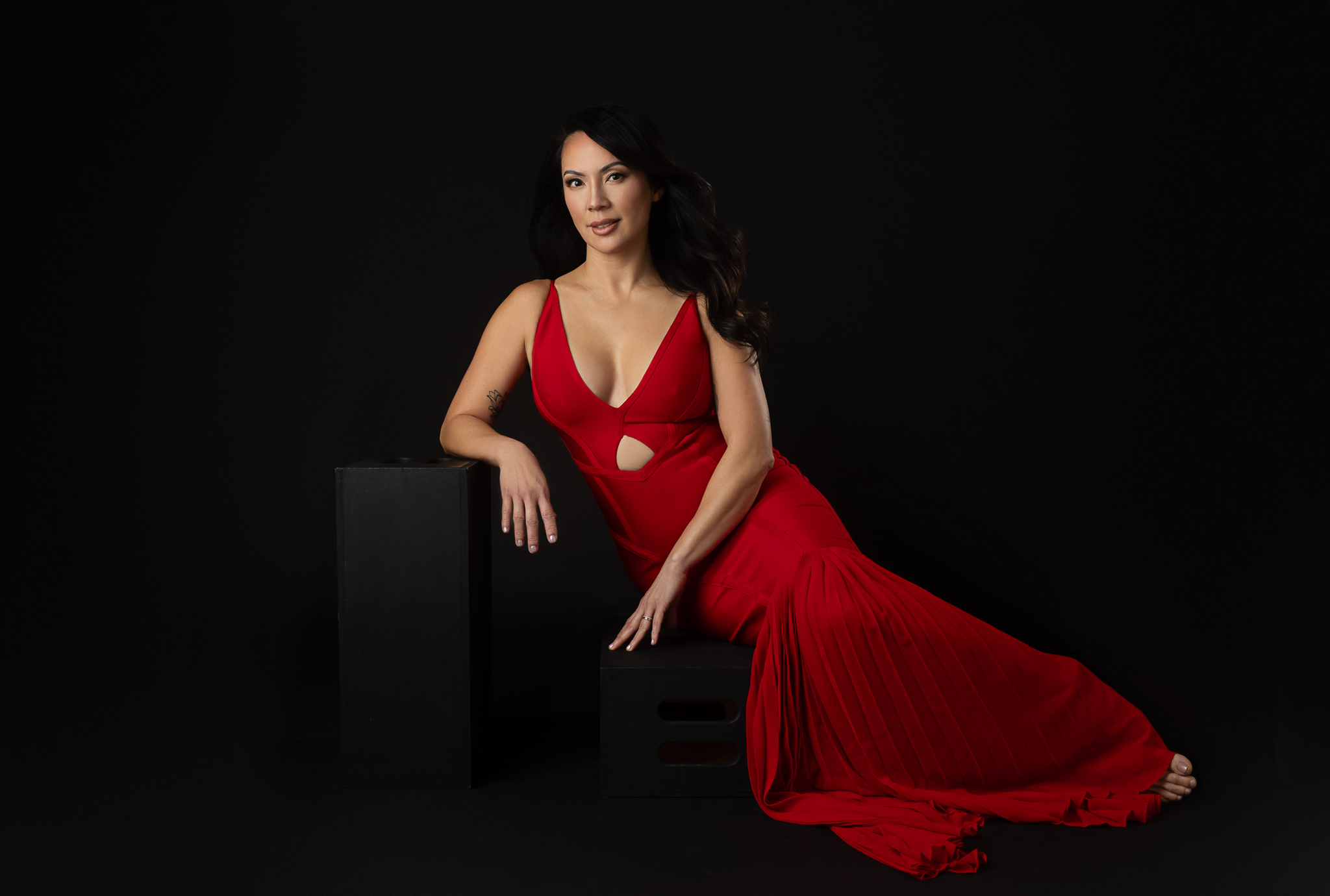 A woman in a red gown sits on a black box in a studio