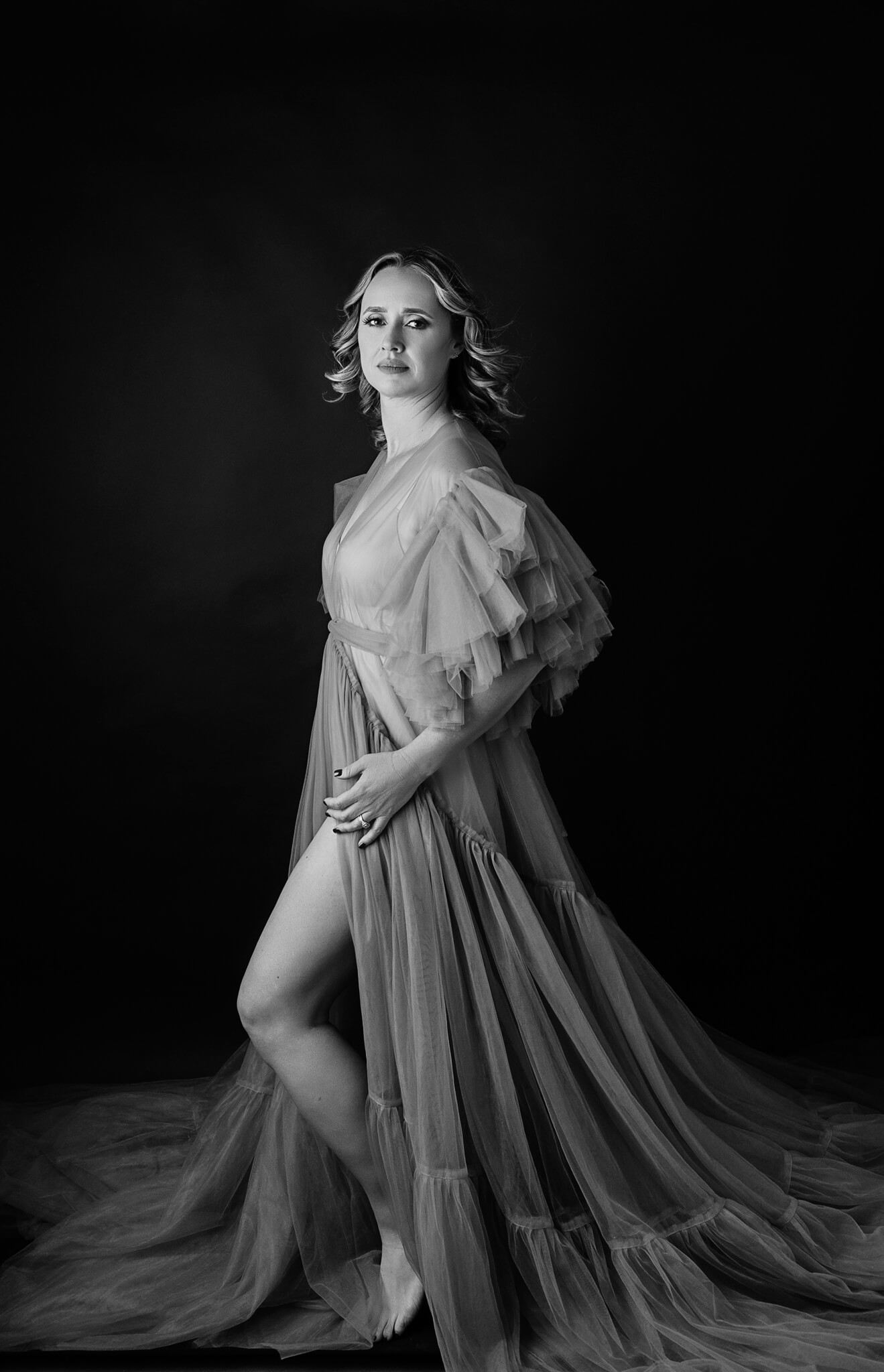 A woman in a long sheer gown stands in a studio with her leg popped out