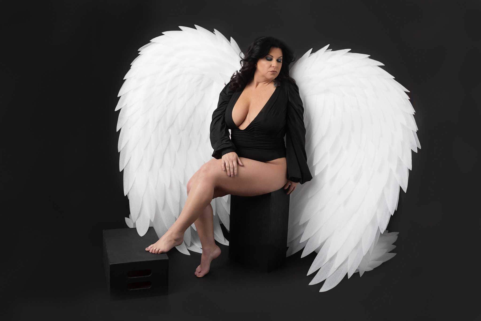 A woman in a black top sits on a box in a studio wearing large white angel wings after visiting waxing studio austin
