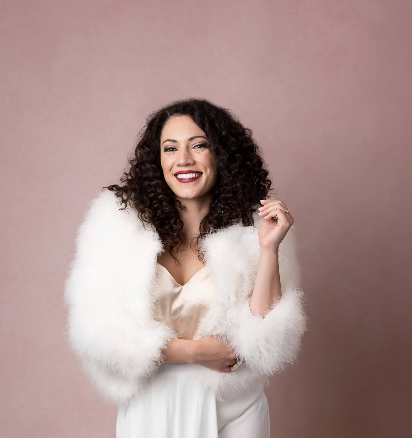 A happy woman with curly dark hair stands in a studio in white pants and a fur sweater