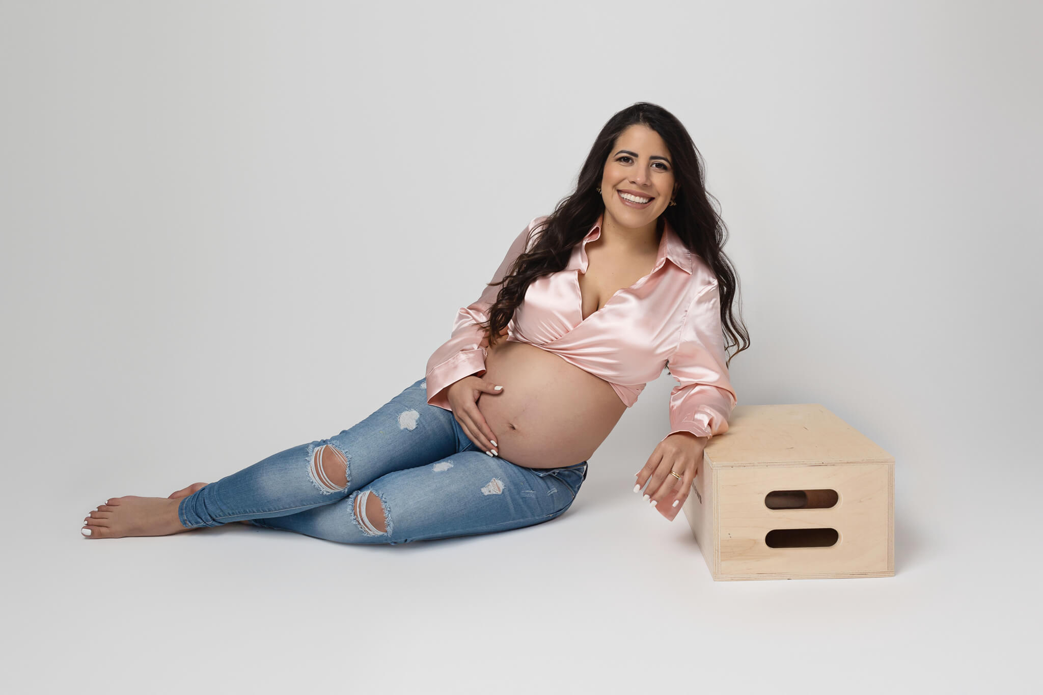 A happy mother to be in a pink top and jeans sits on the floor of a studio holding her bump and leaning on an apple box