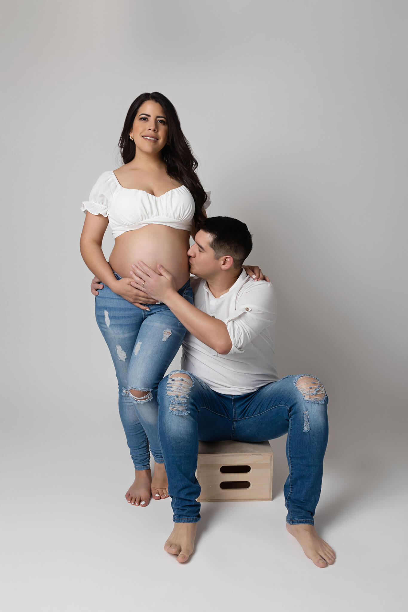 A husband sits on an apple box in a studio kissing the belly of his pregnant wife in jeans and white shirts thanks to fertility clinic austin