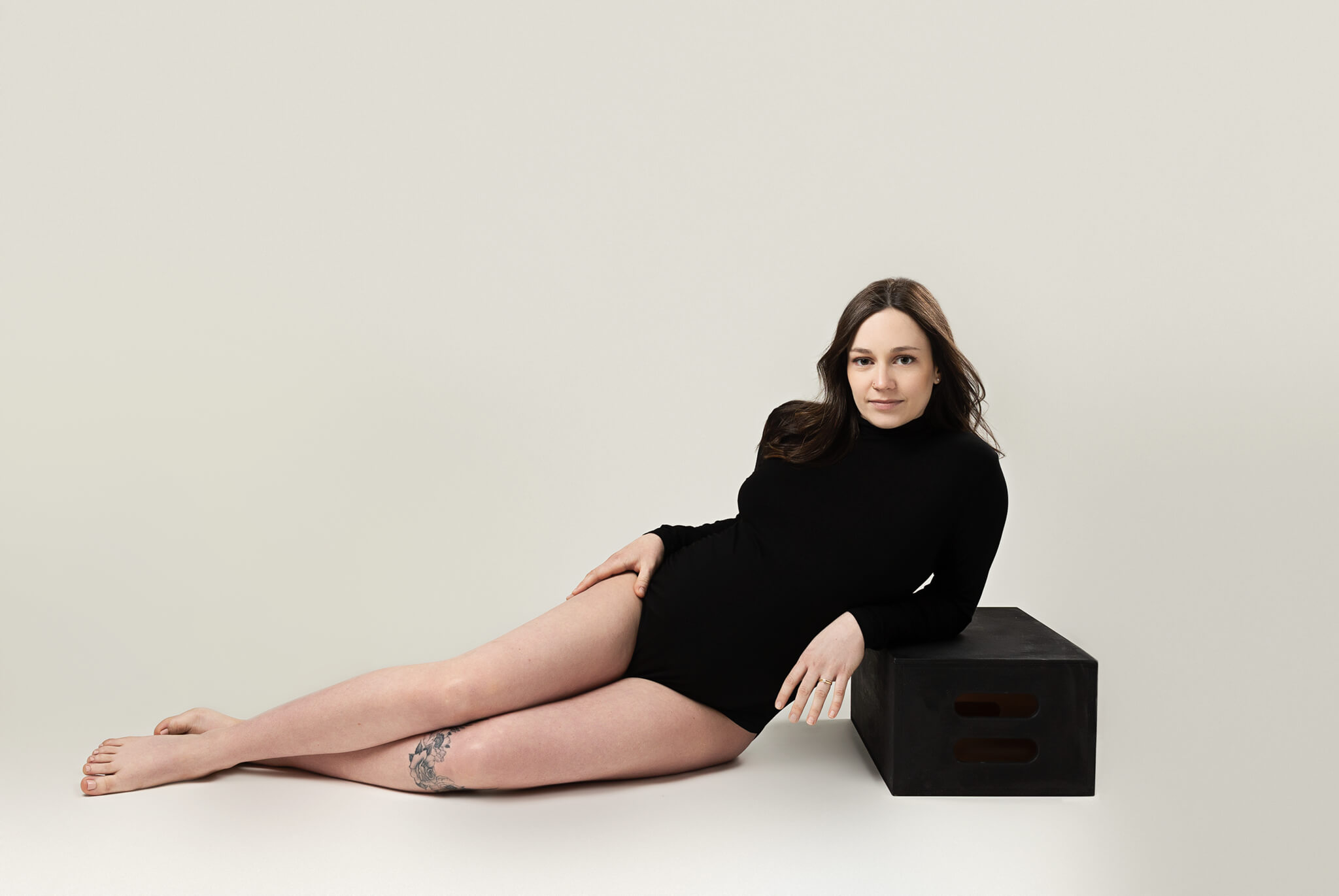 A pregnant woman in a black one-piece lays across a studio floor leaning on a black apple box