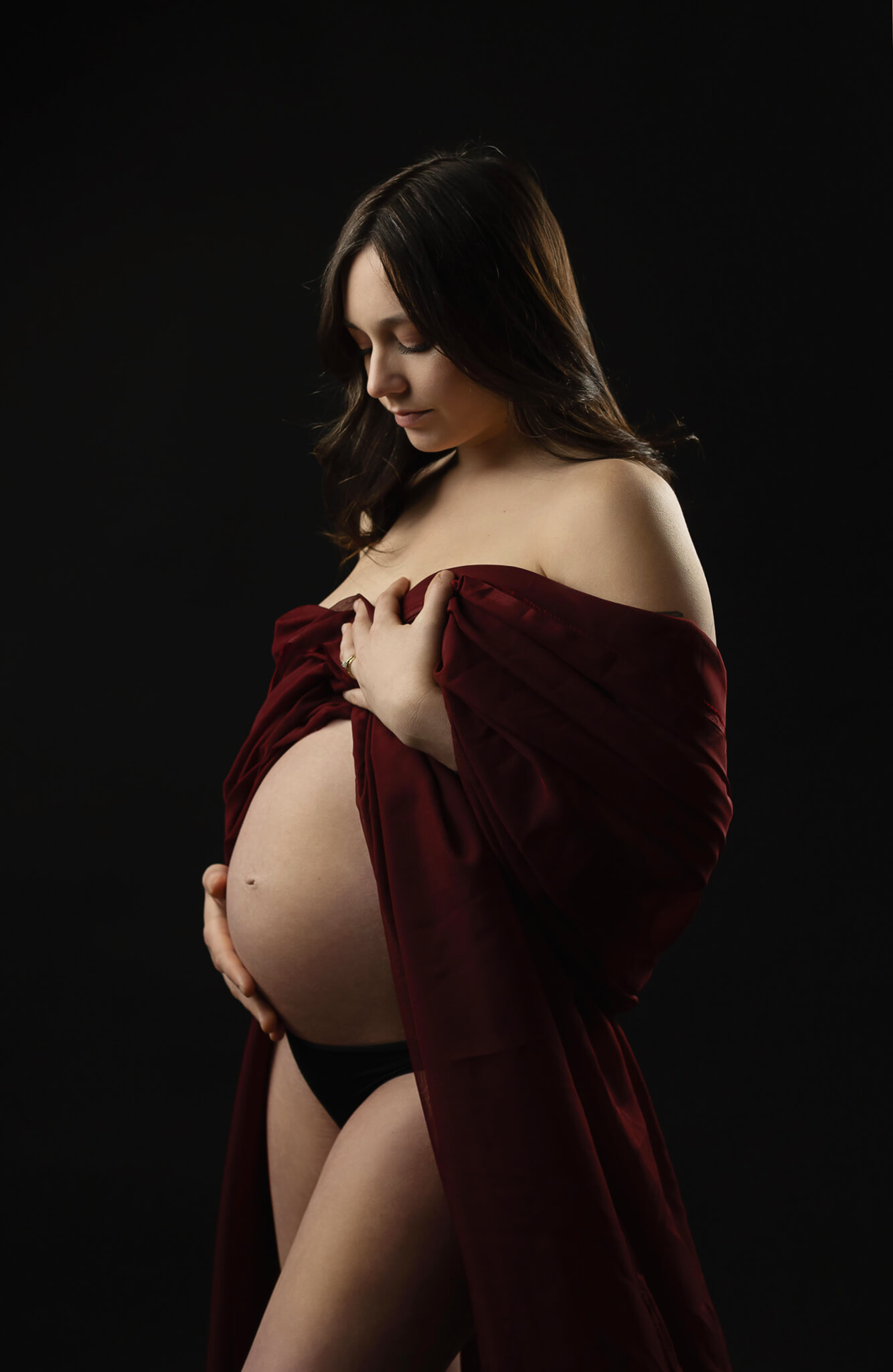 A mother to be draped in a maroon sheet stands in a studio smiling down at her bump after meeting a postpartum doula austin