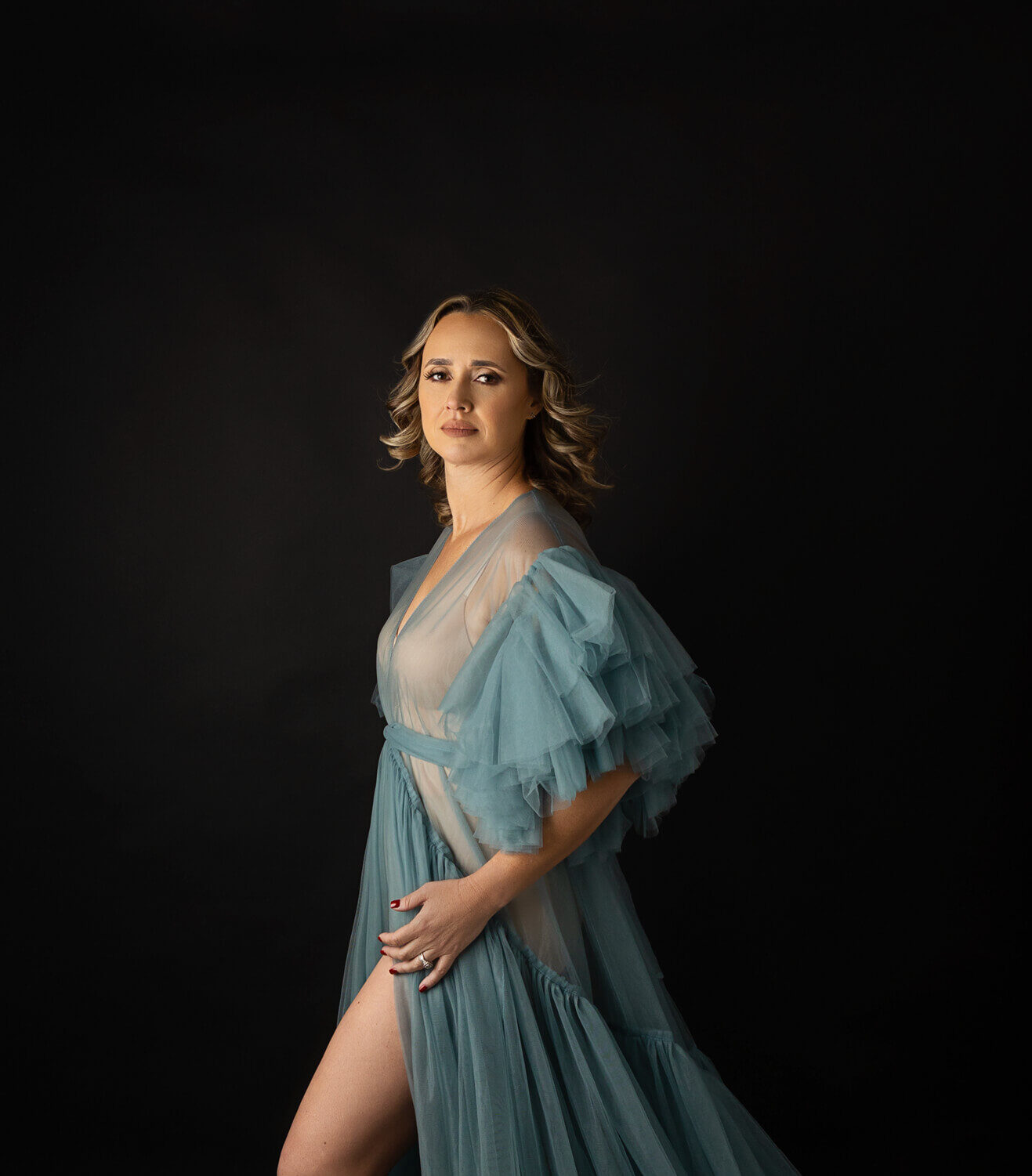 A woman in a long blue tule dress stands in a black studio with a leg popped after using a tanning salon austin