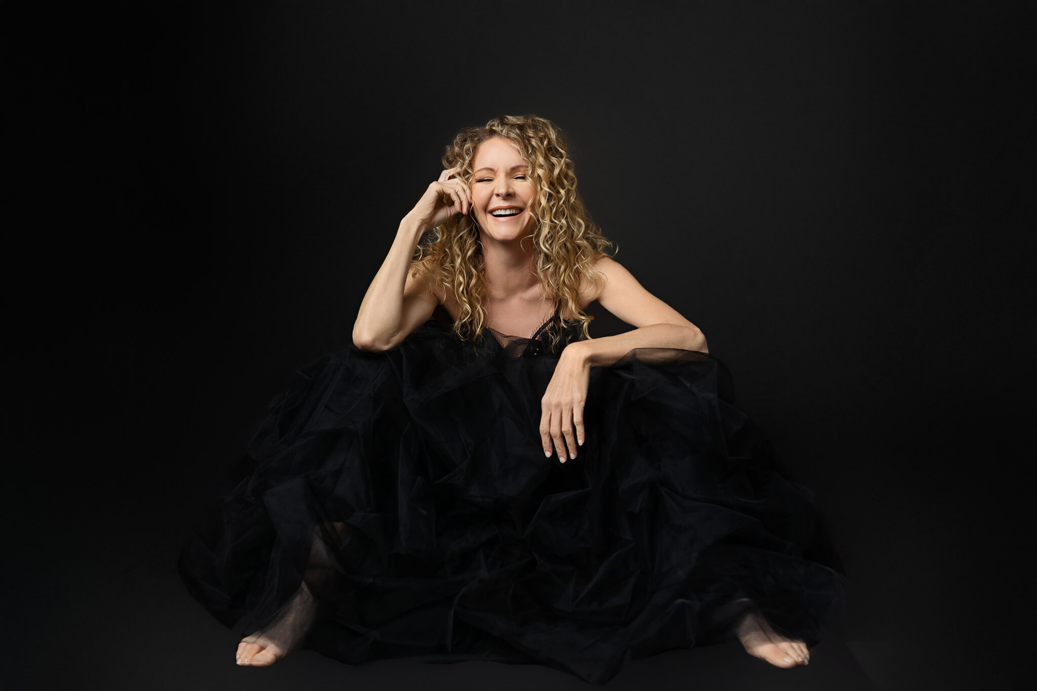 Blonde woman is laughing in a relaxed pose for her photoshoot with a black dress and black backdrop