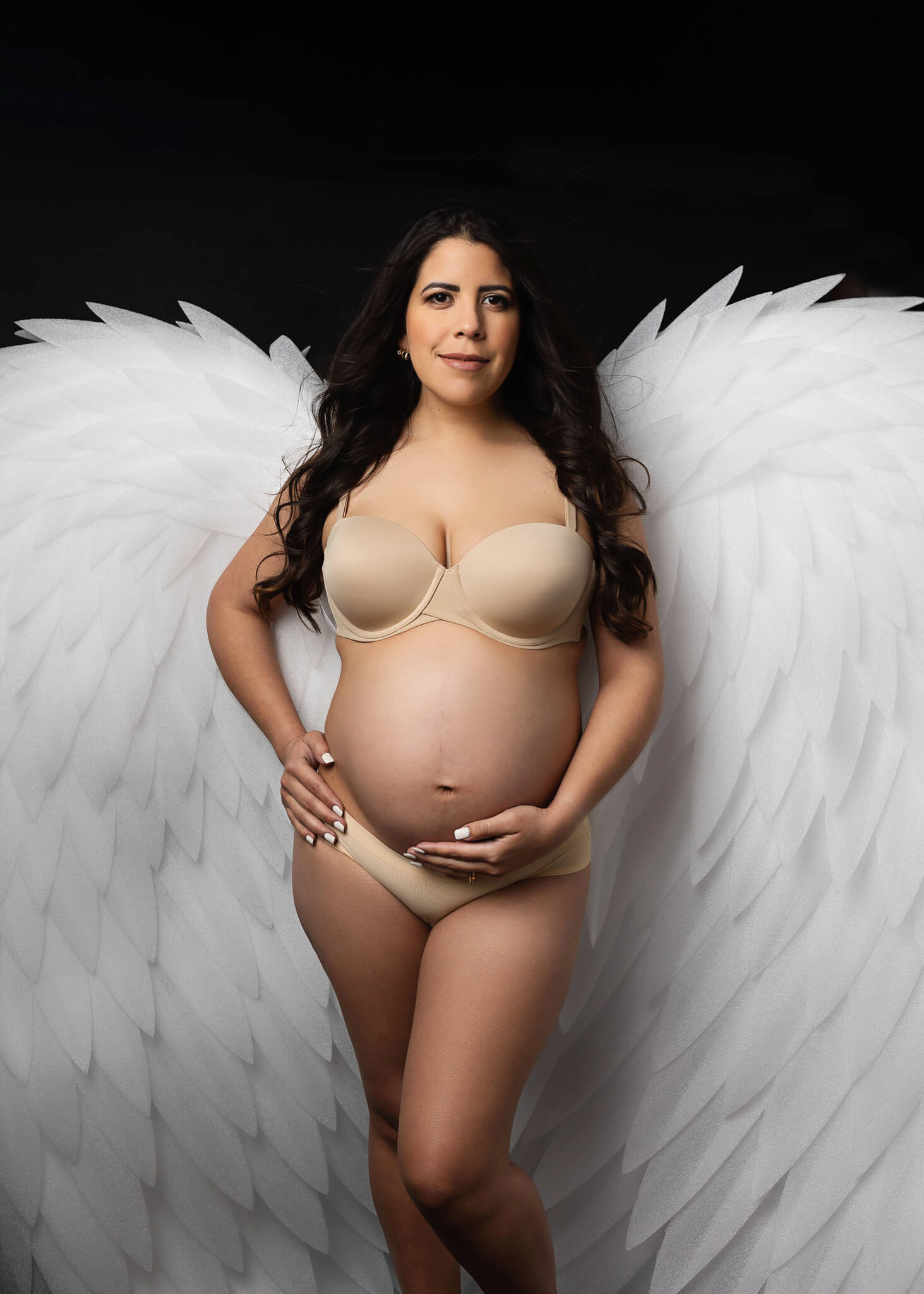 maternity photoshoot of a young woman wearing white wings while holding her belly at Austin studio