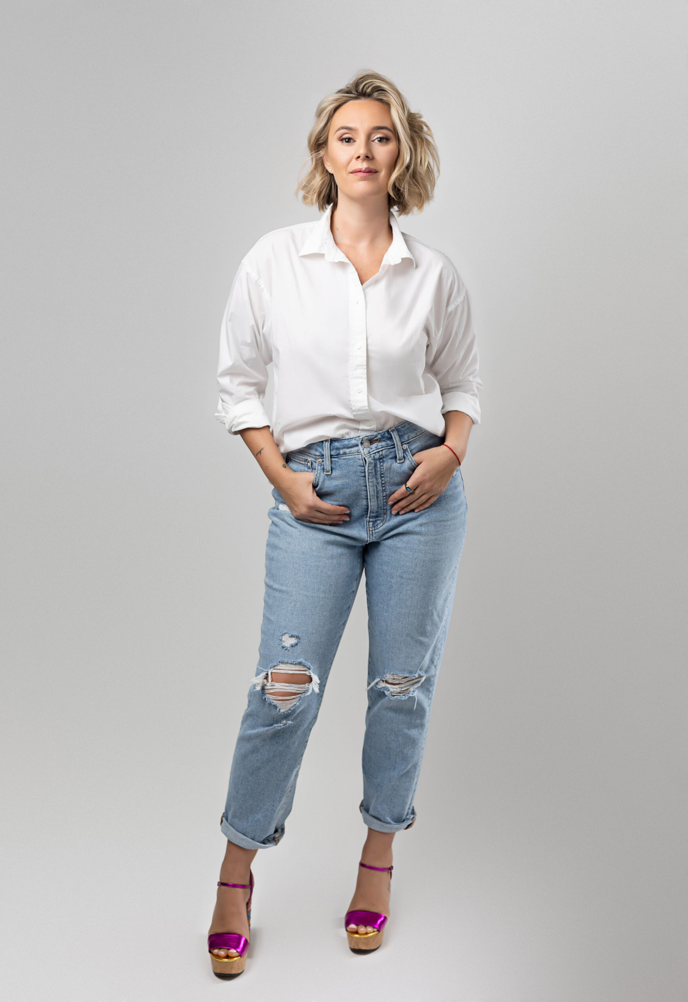 Studio image of a blonde woman wearing jeans and a white shirt over white backdrop with her hand in pockets