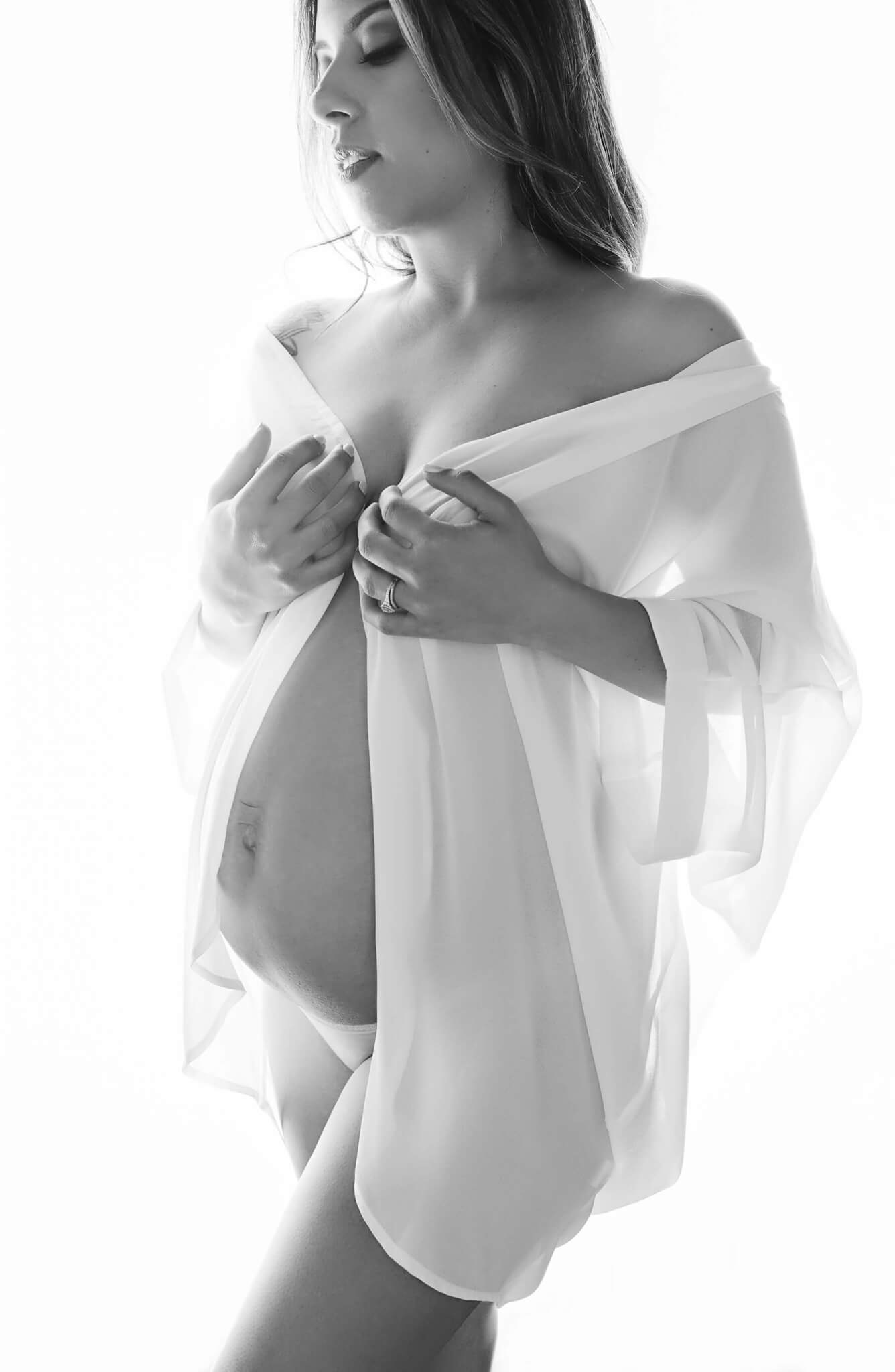 black and white image of a pregnant woman looking down and wearing a white shirt while she shows her belly