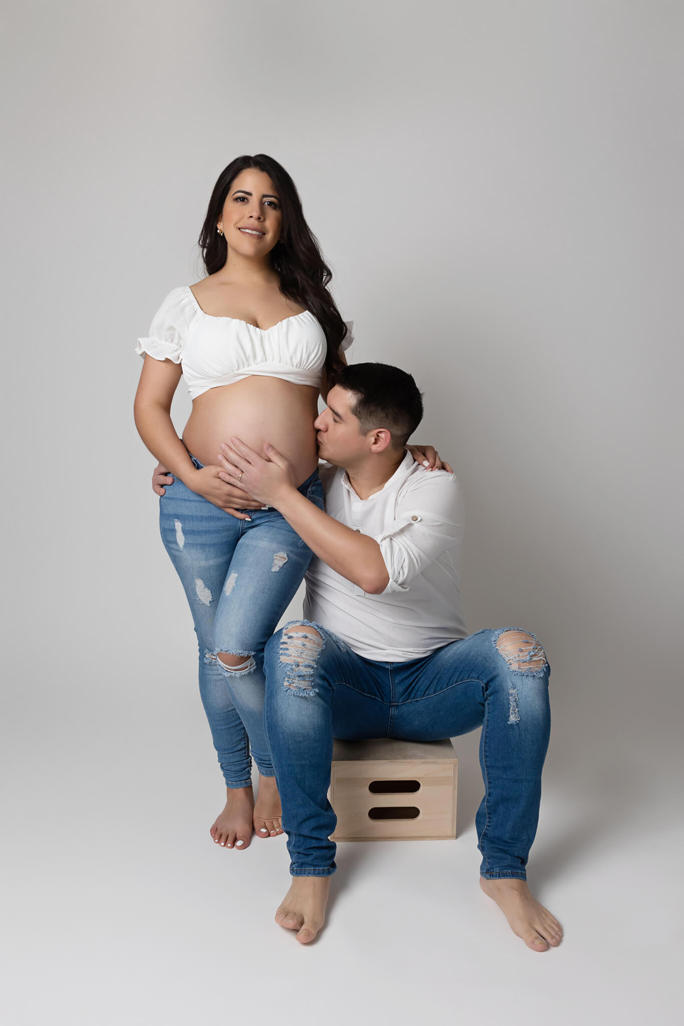 Happy expecting couple are posing, she is standing p and he is sitting and kissing her belly, both are wearing jeans