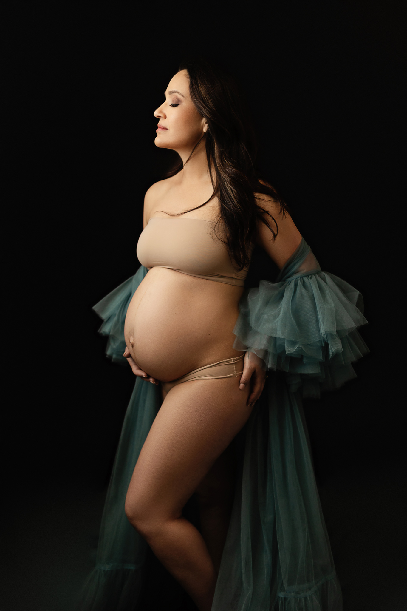 romantic color image of a pregnant woman wearing a big tulle gown in aqua over black backdrop while showing her cute pregnant belly