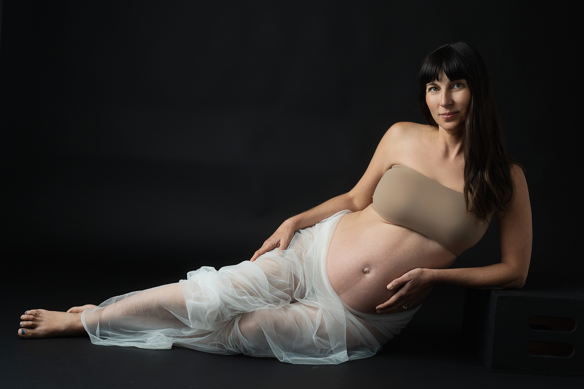dark hair woman is getting a maternity session done white wearing just a tulle fabric and a strapless bra in a very artistic pose
