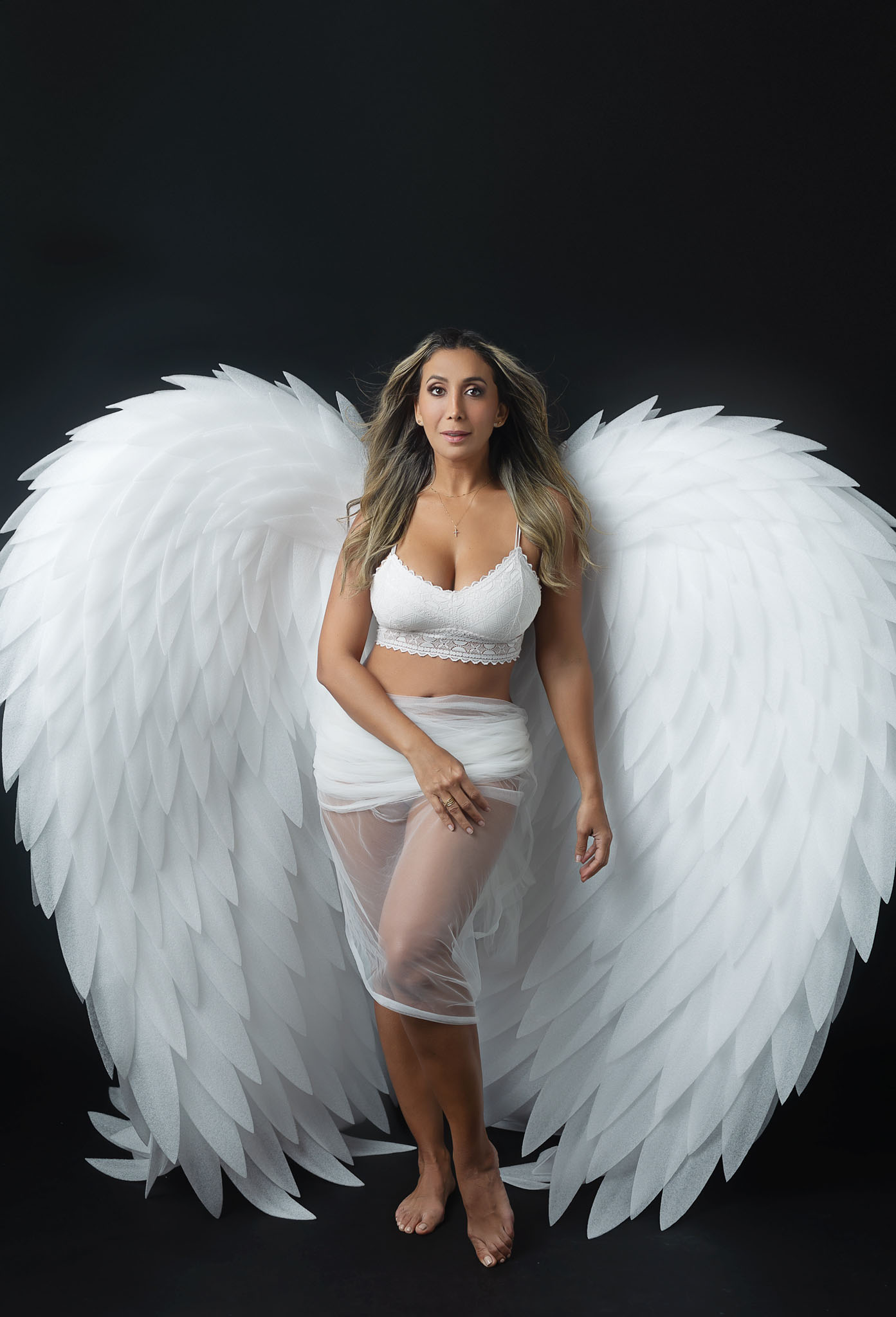 woman in her 40's is posing for her boudoir session, she is wearing white angel wings and white lingerie in a studio setup on black backdrop