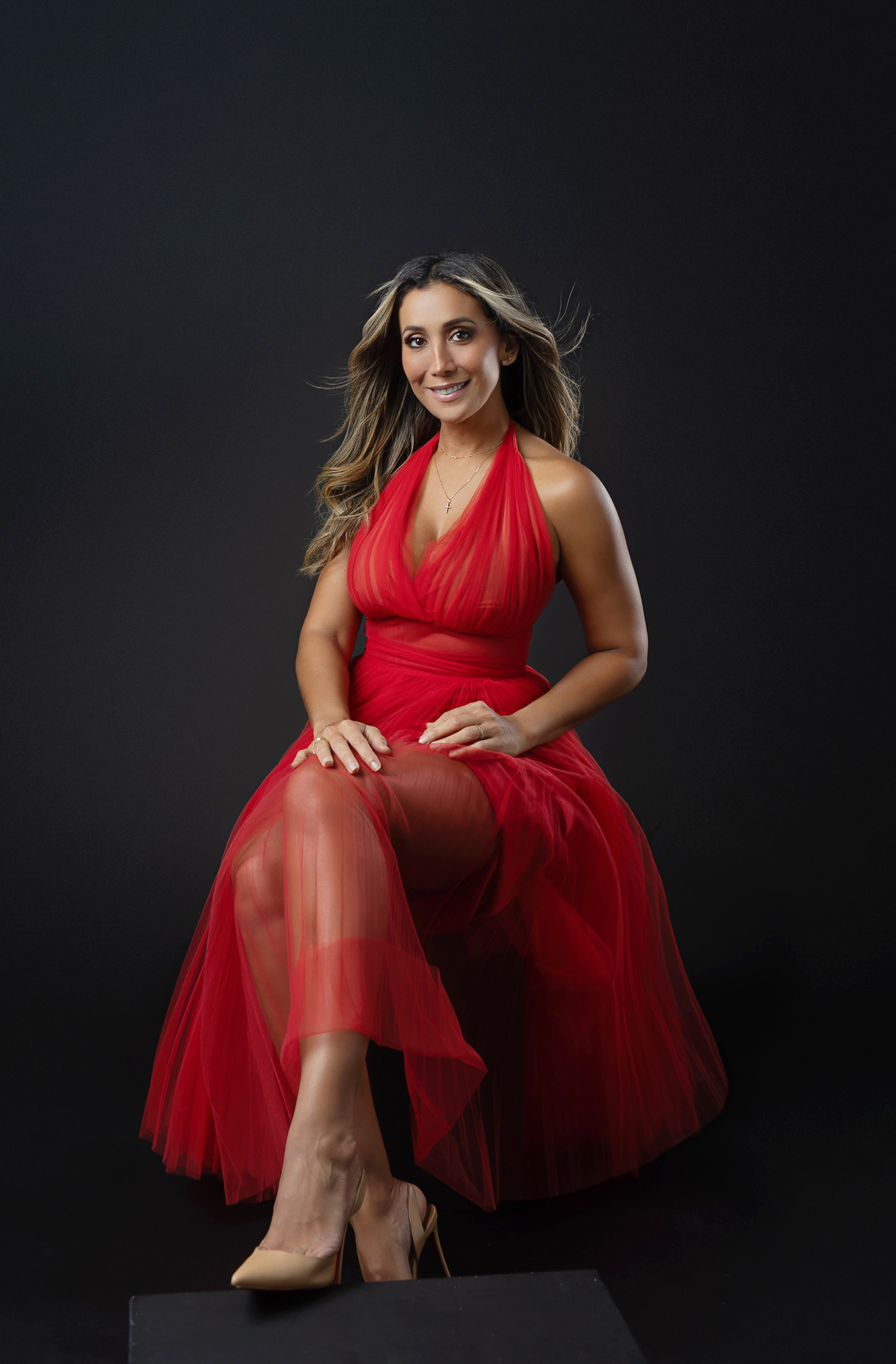 latino woman pose with a tulle red gown on a seated pose while looking very relaxed and comfortable in a studio setting