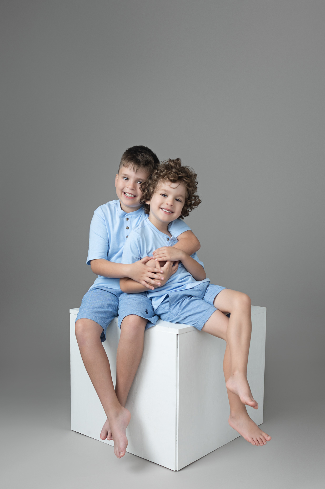 brother boys wearing light blue on a grey backdrop show their big smiles while show affection to each other in Austin studios